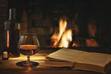 Glass of cognac, a bottle and an open old book on the table near the burning fireplace. Rest and relaxation concept