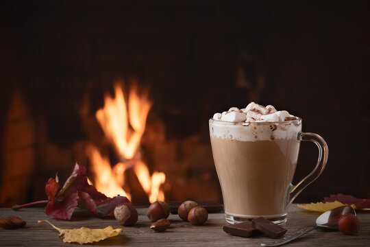 Cocoa with marshmallows and chocolate in a glass mug on a wooden table near a burning fireplace