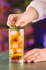 Female bartender putting mint in glass of tasty Cuba Libre cocktail on table in bar