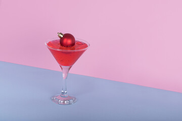 Cocktail glass with red cocktail and Christmas bauble ball on blue and pink background. Minimal Christmas party idea.