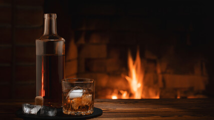 Bottle and glass of whiskey with ice on the table near the burning fireplace. Rest and relaxation...