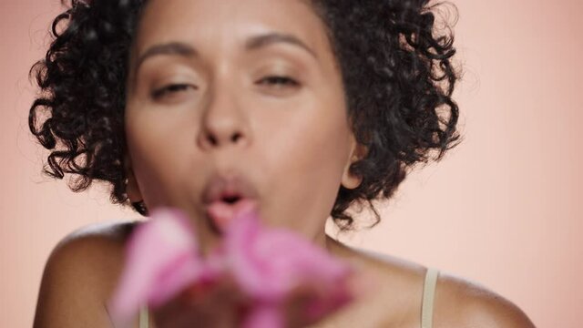 Female Beauty Portrait. Smiling Multiethnic Black Woman Giving a Flying Kiss and Blowing Pink Flower Petals from Her Palms. Wellness, Health, Spa and Natural Skincare Concept on Isolated Background.