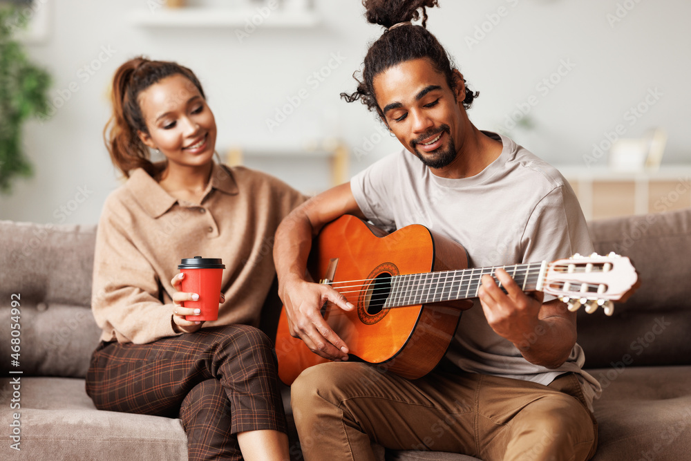 Wall mural young smiling african american man musician playing instumental music on guitar for happy girlfriend