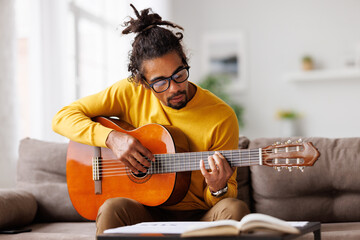 Young joyful african american man playing acoustic guitar at home, sitting on sofa in living room - 468054919