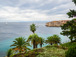 Green palm trees loom over blue Adriatic Sea with sliver of Dubrovnik, Croatia in the background.