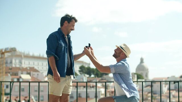 Long shot of happy gay kneeling and proposing to boyfriend. Vertical motion of excited homosexual man opening box with engagement ring in front of lover on background of cityscape. LGBT, love concept
