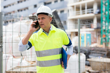 Young confident engineer working at a construction site is talking on a mobile phone, discussing...