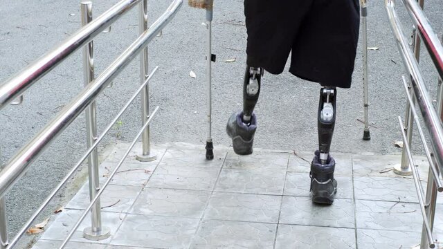 A man living with a disability with amputated legs and arms walks on modern artificial bionic legs prosthesis using old plastic arms tied to crutches with sticky tape