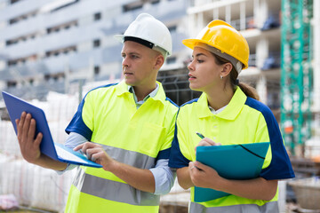 Two builders in uniform planning their work in construction plant. Man using laptop, woman holding paper folder in hands.