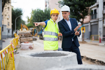 Male architect in suit and white hardhat and woman builder in uniform standing and discussing their work while standing in construction area. Woman pointing with finger.