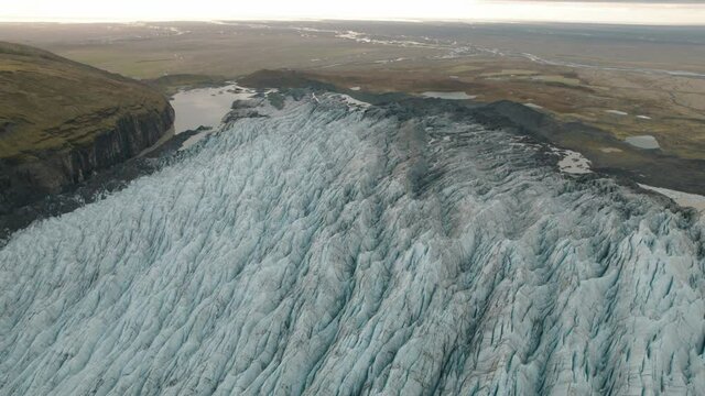 Large glacier in Iceland flowing down mountain to green plains. Aerial tilt up.