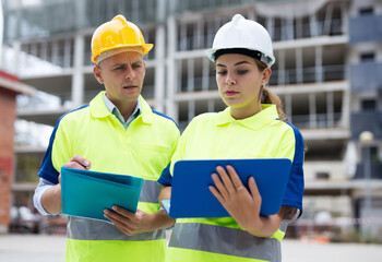 Two engineers working on a construction site are holding a laptop and an estimate in their hands,...