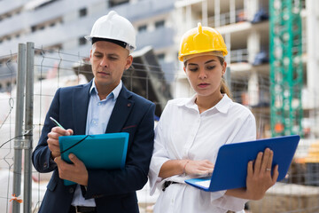 Male and female egineers standing in construction site and planning project of new multistory building. Woman holding laptop and man holding paper-folder in hands.