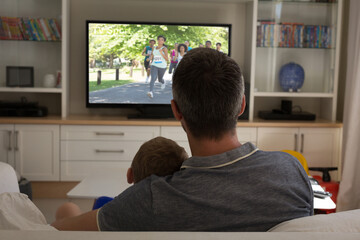 Rear view of father and son sitting at home together watching athletics event on tv