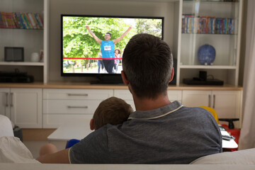 Rear view of father and son sitting at home together watching athletics event on tv