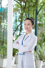 Portrait of positive young Asian physician crossing arms when standing at big window in patients house