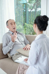 Senior man sitting on sofa and talking to psychotherapist taking notes in medical card