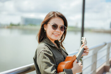 Obraz na płótnie Canvas Playing Ukulele of Young Beautiful Asian Woman Wearing Jacket And Black Jeans Posing Outdoors