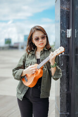 Obraz na płótnie Canvas Playing Ukulele of Young Beautiful Asian Woman Wearing Jacket And Black Jeans Posing Outdoors