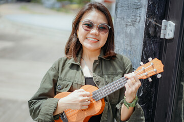 Playing Ukulele of Young Beautiful Asian Woman Wearing Jacket And Black Jeans Posing Outdoors