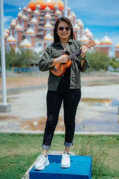 Playing Ukulele of Young Beautiful Asian Woman Wearing Jacket And Black Jeans Posing Outdoors