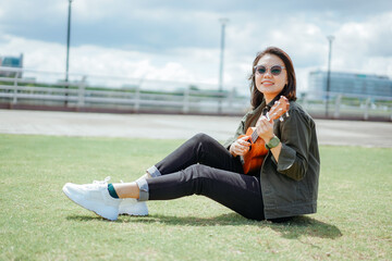Playing Ukulele of Young Beautiful Asian Woman Wearing Jacket And Black Jeans Posing Outdoors