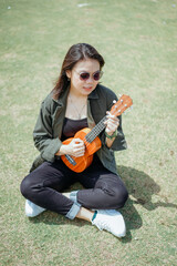 Playing Ukulele of Young Beautiful Asian Woman Wearing Jacket And Black Jeans Posing Outdoors