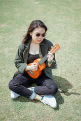 Playing Ukulele of Young Beautiful Asian Woman Wearing Jacket And Black Jeans Posing Outdoors