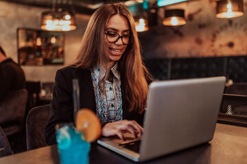 Business woman in glasses sitting in a modern cafe and working on her laptop while drinking a cocktail. Selective focus