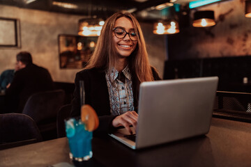 Fototapeta premium Business woman in glasses sitting in a modern cafe and working on her laptop while drinking a cocktail. Selective focus