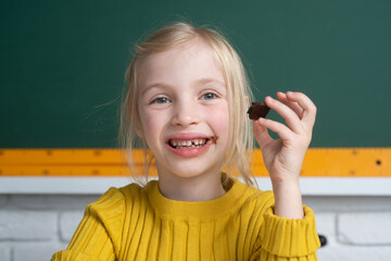 Close-up portrait of smiling little happy girl sitting on table desktop in class room, eating...