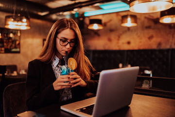 Business woman in glasses sitting in a modern cafe and working on her laptop while drinking a cocktail. Selective focus