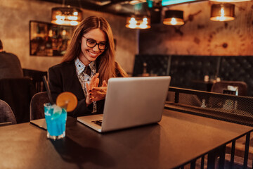 Business woman in glasses sitting in a modern cafe and working on her laptop while drinking a cocktail. Selective focus