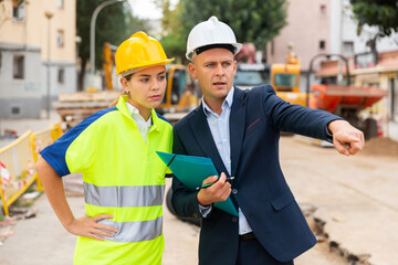 Two confident engineers in hard hats discussing blueprint while standing at construction site
