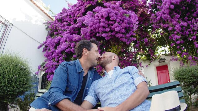 Happy gay couple sitting on bench in park and doing selfie. Front view of smiling homosexual male lovers looking at camera, spending weekend together in park with blooming trees. LGBT, date concept