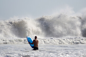 Fototapeta premium A blurred unrecognizable man's figure with a bodyboard in front of a big wave braking on a beach, vertical orientation, focus on the wave