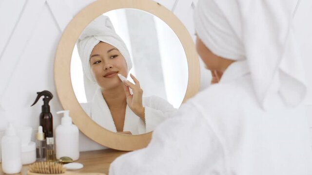 Skin cleansing procedures. Young asian lady cleaning her face with lotion on cotton pad, looking at mirror after shower