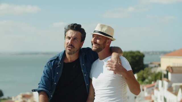 Happy homosexual man sitting and hugging lovers shoulder. Front view of cheerful gay couple having romantic date at observation tower, enjoying time spending together and talking. LGBT, dating concept