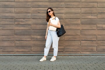 Photo of fascinating beautiful smiling charming young brunette girl wearing stylish clothes holding computer laptop and black sunglasses in white t-shirt and light blue jeans with black bag in the