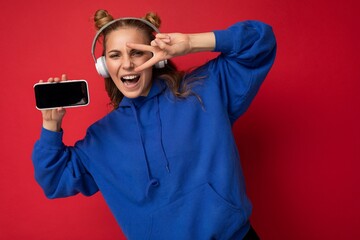 Photo of beautiful positive happy funny young woman wearing stylish blue hoodie isolated over red background with free space for text wearing white bluetooth wireless headphones and listening to music