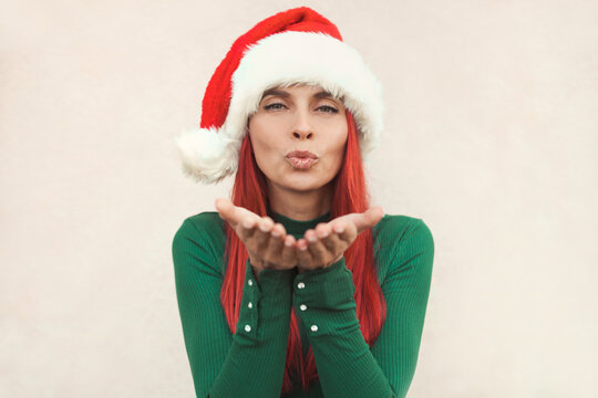 Close up emotional portrait of happy smiling hipster woman 30-35 years old with long red hair doing air kiss, wearing green sweater and santa claus hat against light wall background