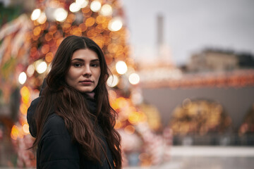 A beautiful and young girl looks into the camera. Christmas city decoration blurred in the background. Cute brunette faced to the camera in the downtown before New Year party