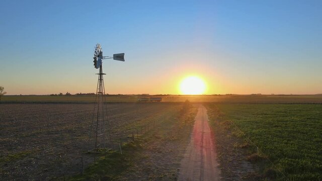 Cinematic aerial pan shot capturing old school rustic windmil on agriculture farmland with pick up truck driving pass in the background at beautiful sunset golden hours, La Pampa, Patagonia.