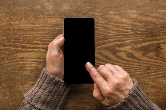 Old Mature Female Hands Holding Smartphone And Touching Black Screen With Finger On Brown Wooden Table Background. Senior Woman Using Modern Device. Closeup. Empty Place For Text. Top Down View.