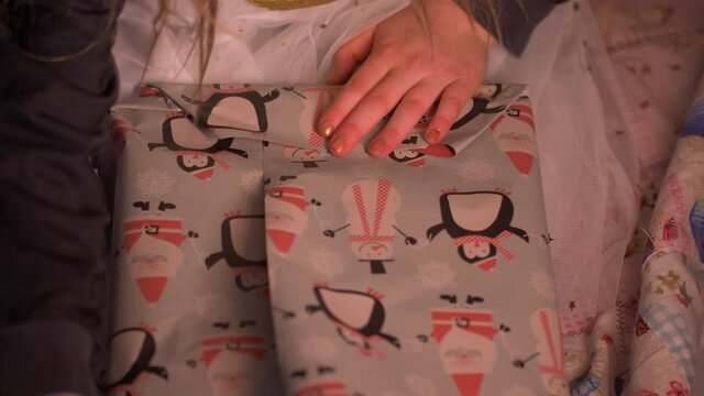 Close-up of little girl wrapping birthday presents or Christmas gift at home. Frontal View of female child hands decorating presents with grey craft paper with penguins using tape. 4K Pro Res.