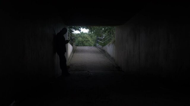 Silhouette Of Adult Male In Underpass Tunnel Checking Phone And Leaning Against Wall Taking Phone Call. Static, Establishing Shot