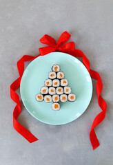 Christmas tree made of sushi on blue round plate with red bow and ribbon on a gray background. A creative idea for a Japanese restaurant. Vertical photo. Top view. Imitation of a Christmas ball.