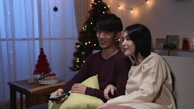 funny couple watching tv sitting on couch in living room at home in winter night during christmas holiday. young asian girl and boy point finger to television screen and laughing talking on sofa