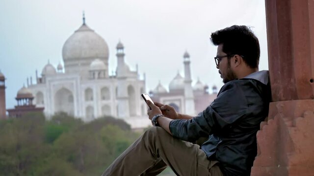 A young man or male in eye glasses sitting against the taj Mahal mausoleum and using a mobile phone or smartphone to type a text message. A freelance professional is browsing on a cellphone 