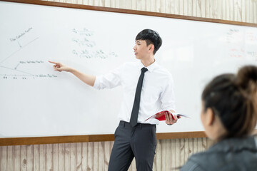 An Asian male teacher in a classroom near the whiteboard is writing and teaching a lesson with a...